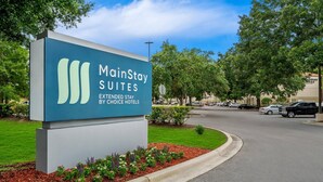 A MainStay Suites sign with a modern design, located at a street corner with a landscaped area and a road in the background.