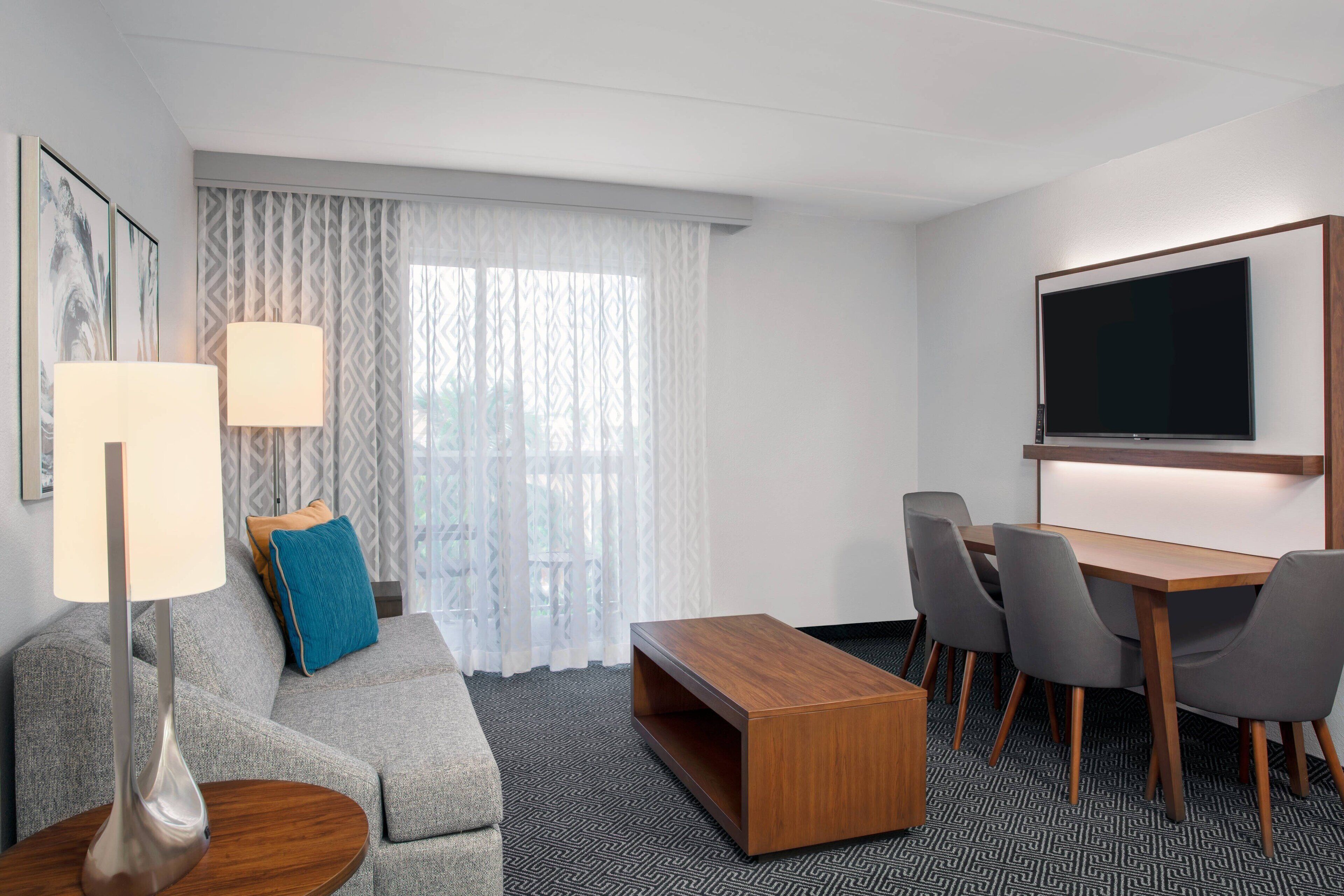 Une chambre d’hôtel avec un canapé gris, une table basse en bois, une télévision et une table à manger avec des chaises.