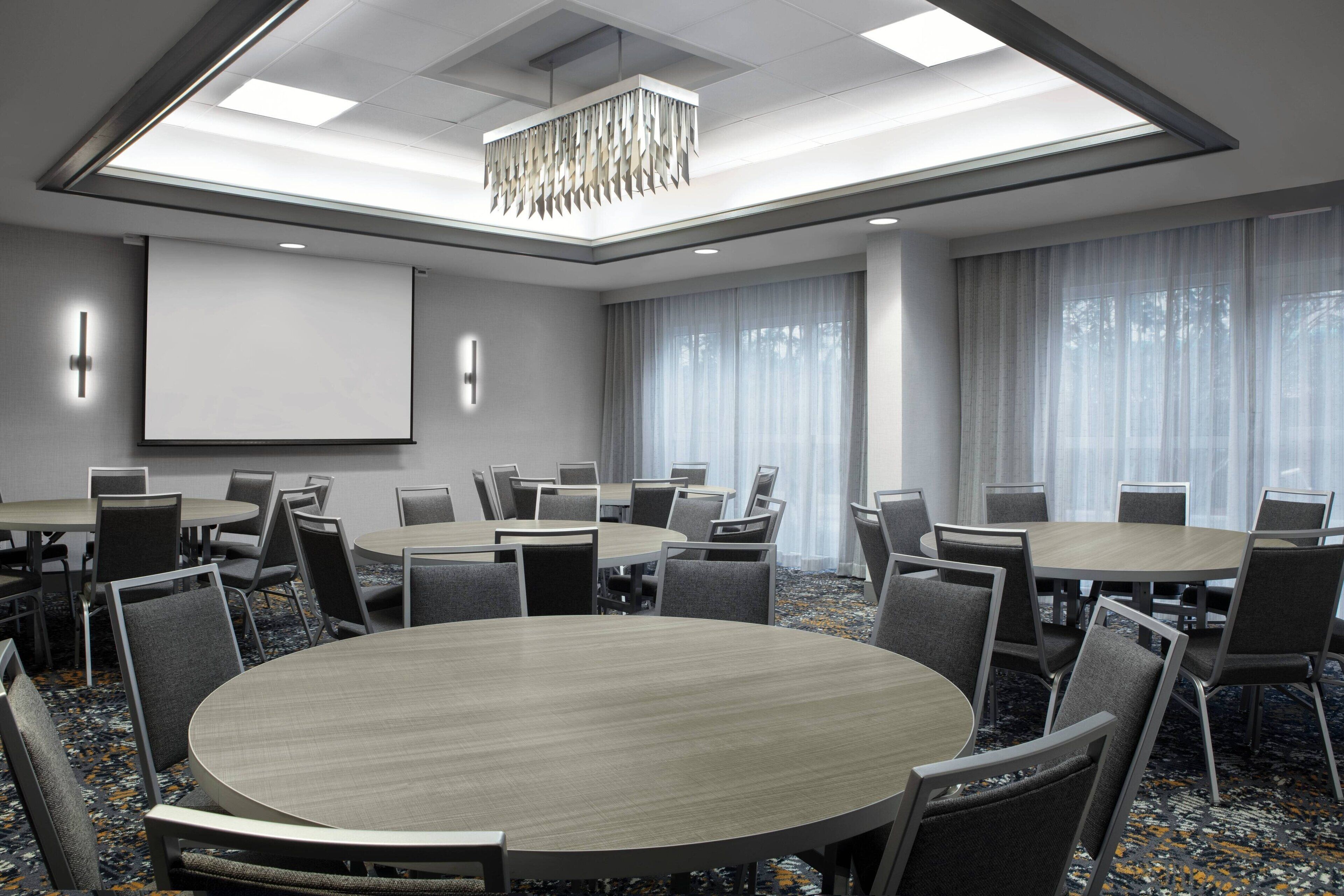 A conference room with multiple tables and chairs, a projector screen, and large windows with curtains.