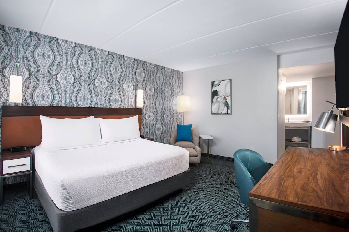 Quarto de hotel com uma cama grande, uma escrivaninha, uma cadeira, uma mesinha de cabeceira e um papel de parede estampado.
