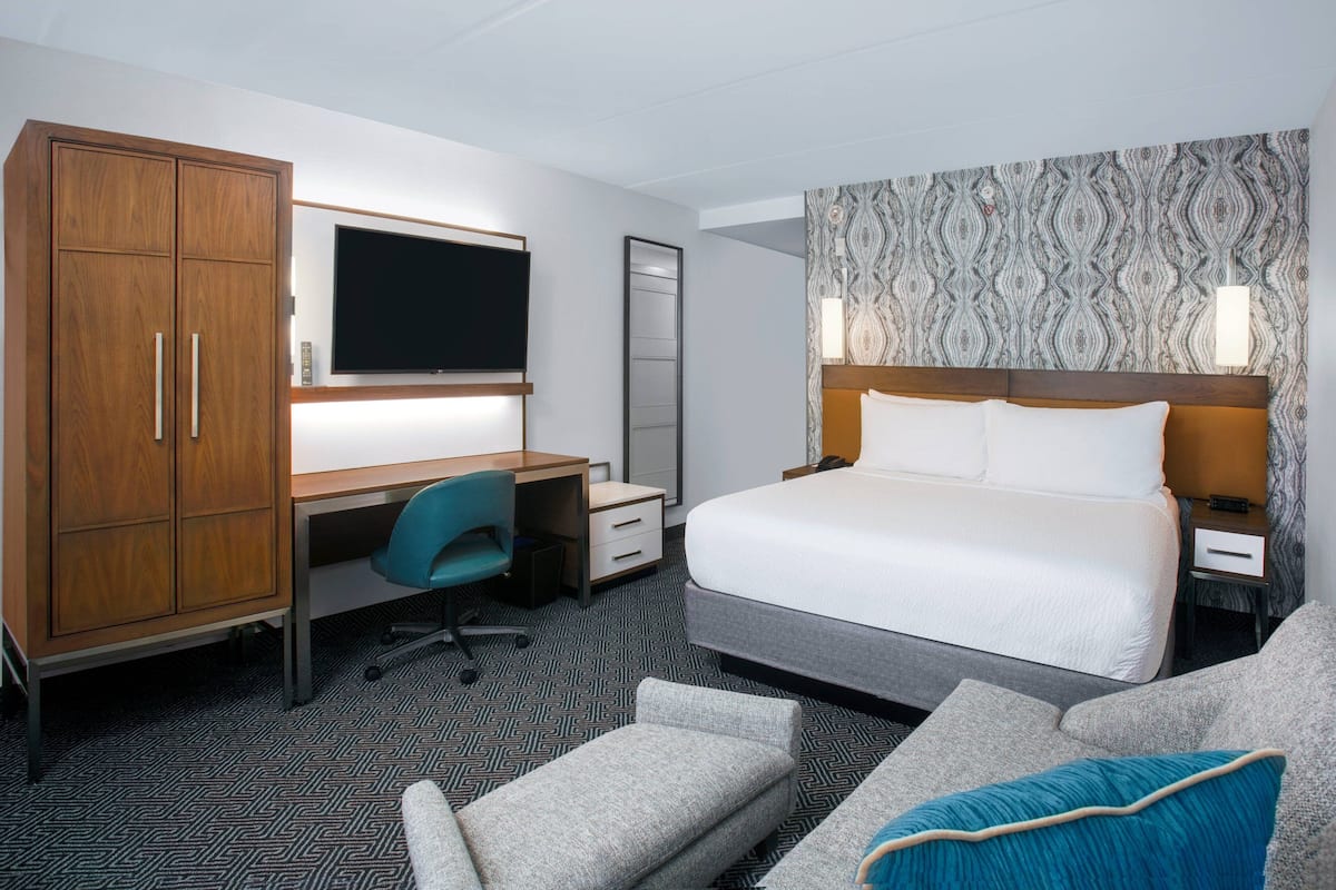 Quarto de hotel com cama, escrivaninha com cadeira, guarda-roupa, televisão e parede estampada.