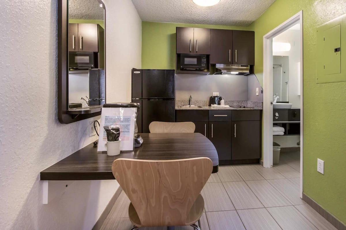 A compact kitchen with dark cabinets, a microwave, and a refrigerator. There is a dining table with a chair and a coffee maker.