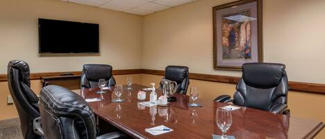 A conference room with a long table, four black leather chairs, a flat-screen TV on the wall, and a framed painting above the table.