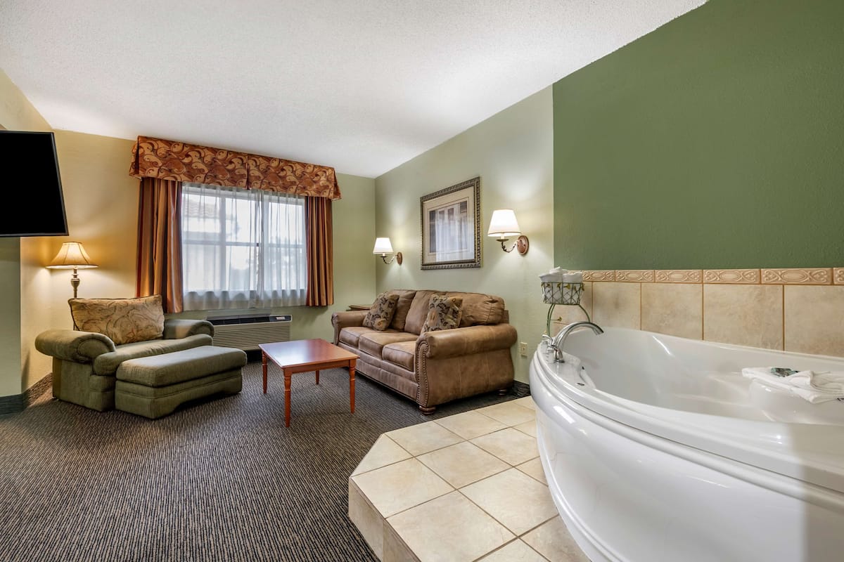 A hotel room with a sofa, a chair, a small table, a large bathtub, and a television.
