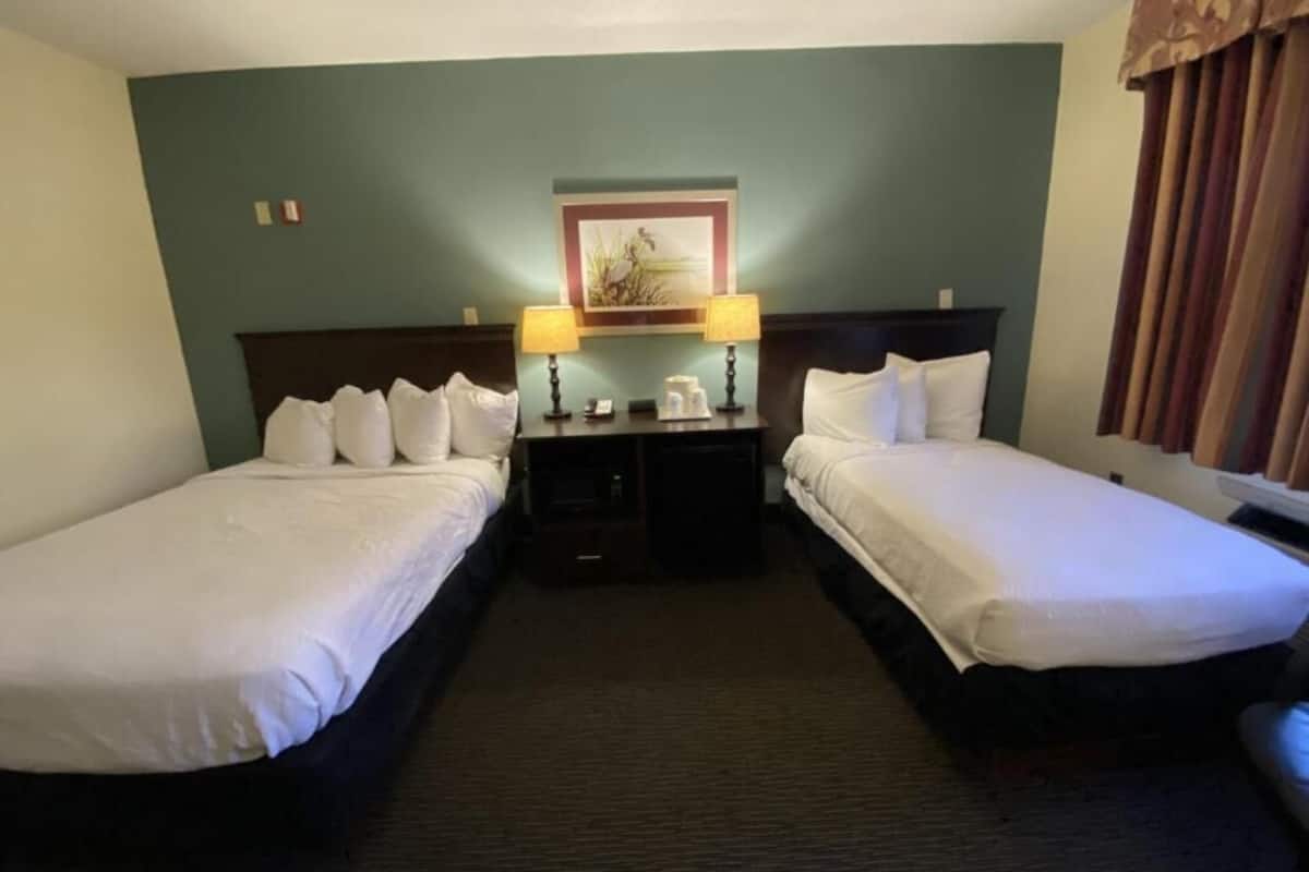 A hotel room with two single beds, a nightstand with a lamp, a small table, and a framed picture on the wall.