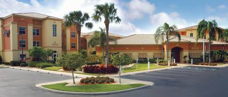 A hotel with a parking lot, palm trees, and a clear sky.