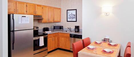 A compact kitchen with wooden cabinets, a stainless steel refrigerator, a microwave, and a coffee maker. There is a dining table with red chairs and a framed picture on the wall.