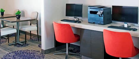 Un espace de travail aménagé dans une chambre d’hôtel, comprenant deux bureaux, chacun équipé d’un écran d’ordinateur, d’un clavier et d’une imprimante. On y trouve également deux chaises rouges et une petite table sur laquelle sont posées des plantes en pot.
