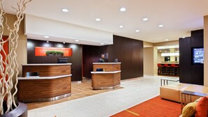 Ein moderner Hotel-Empfangsbereich mit zwei Empfangstresen, einem digitalen Bildschirm und einer Sitzecke mit rotem Teppich.