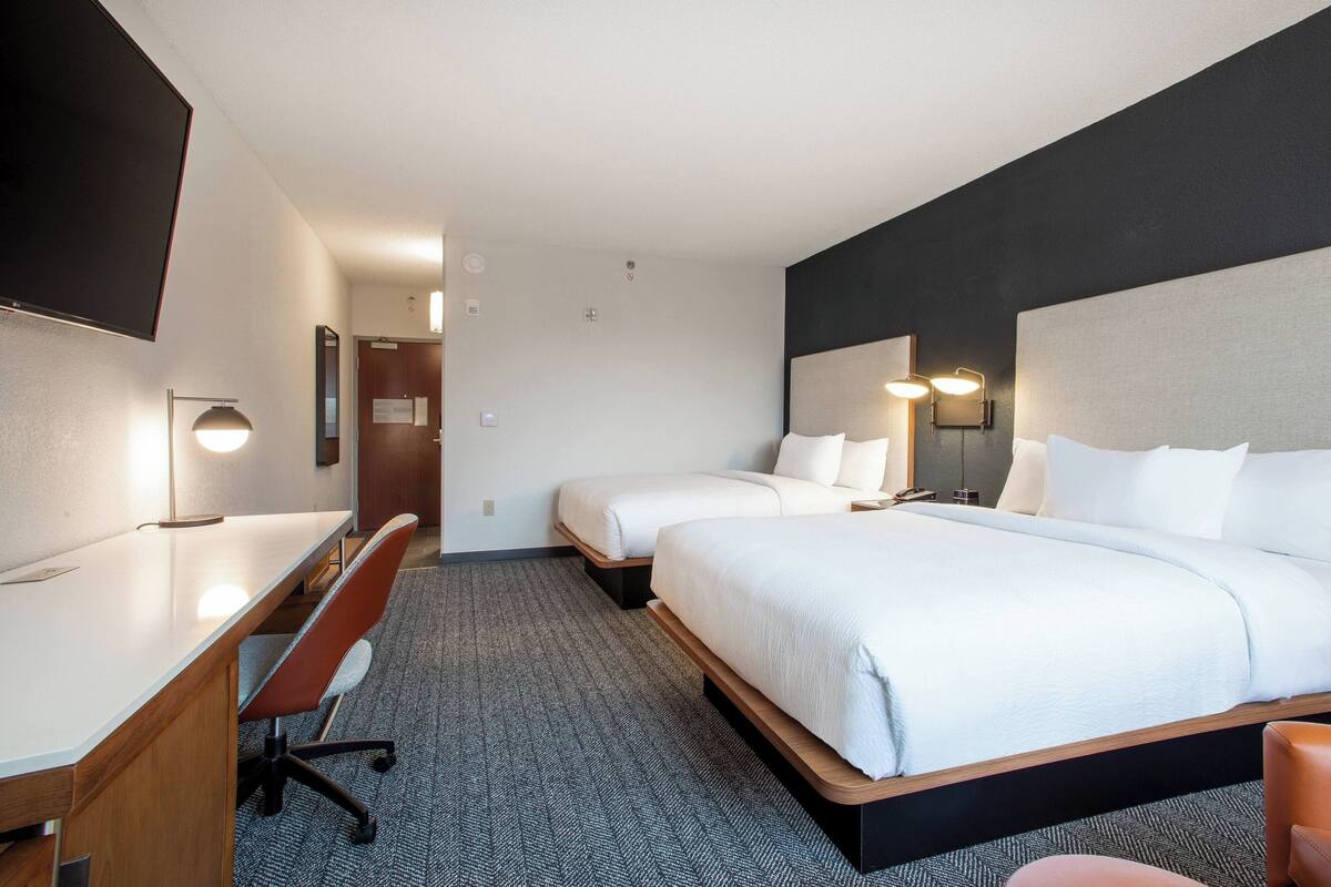 A hotel room with a desk, two beds, a chair, and a TV.