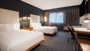 Ein Hotelzimmer mit zwei Betten, einem Schreibtisch, einem Stuhl, einem Fernseher und einem Fenster mit Vorhängen.