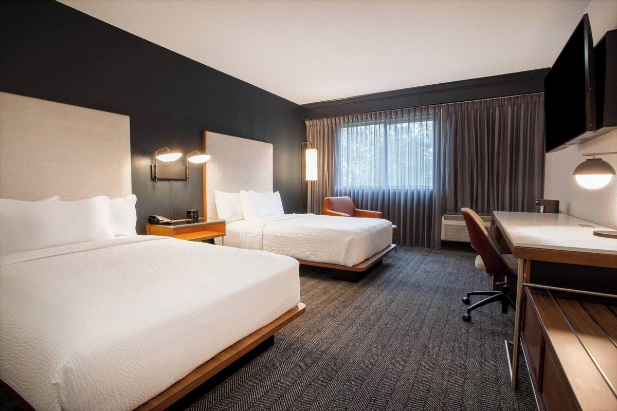 A hotel room with two beds, a desk, a chair, a TV, and a window with curtains.