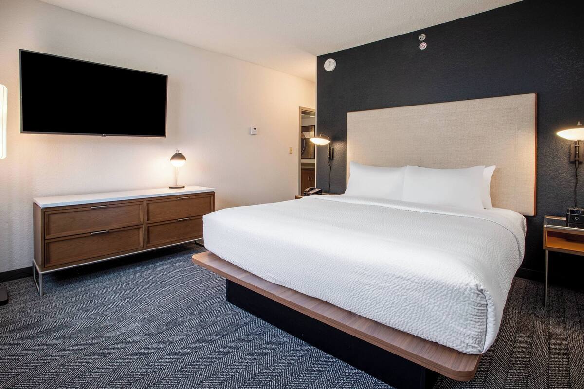 A hotel room with a large bed, a flat-screen TV mounted on the wall, a bedside table with a lamp, and a wooden bench at the foot of the bed.