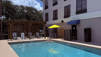 A swimming pool with a metal ladder, a wooden pergola, and a building with multiple windows.