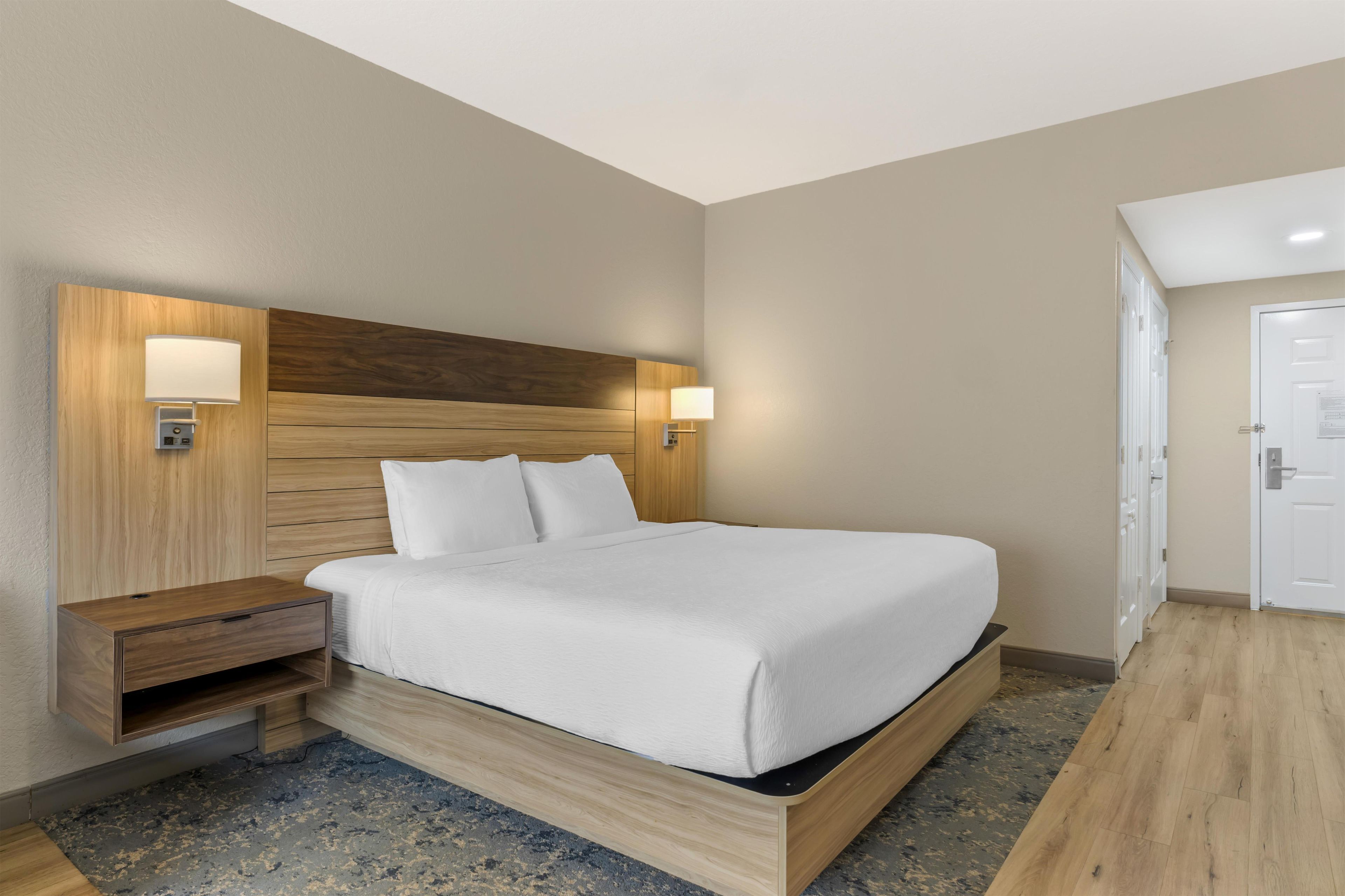 A hotel room with a wooden headboard, a bed with white bedding, a nightstand, and a wall-mounted lamp.