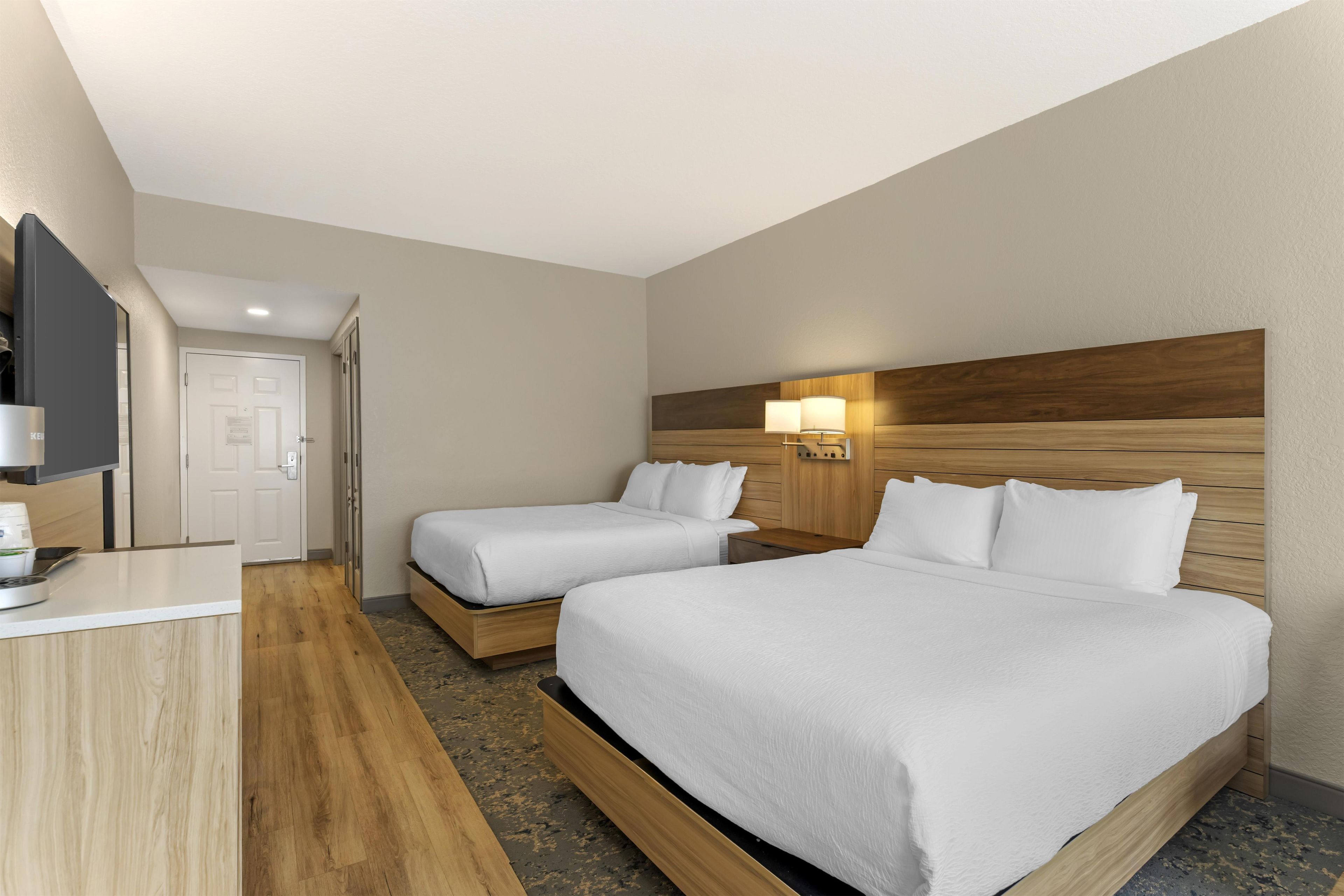 A hotel room with two beds, a desk, a TV, and a wall-mounted lamp.