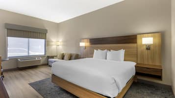 A hotel room with a large bed, two bedside tables, a window with blinds, a wall-mounted heater, and a small table with a chair.