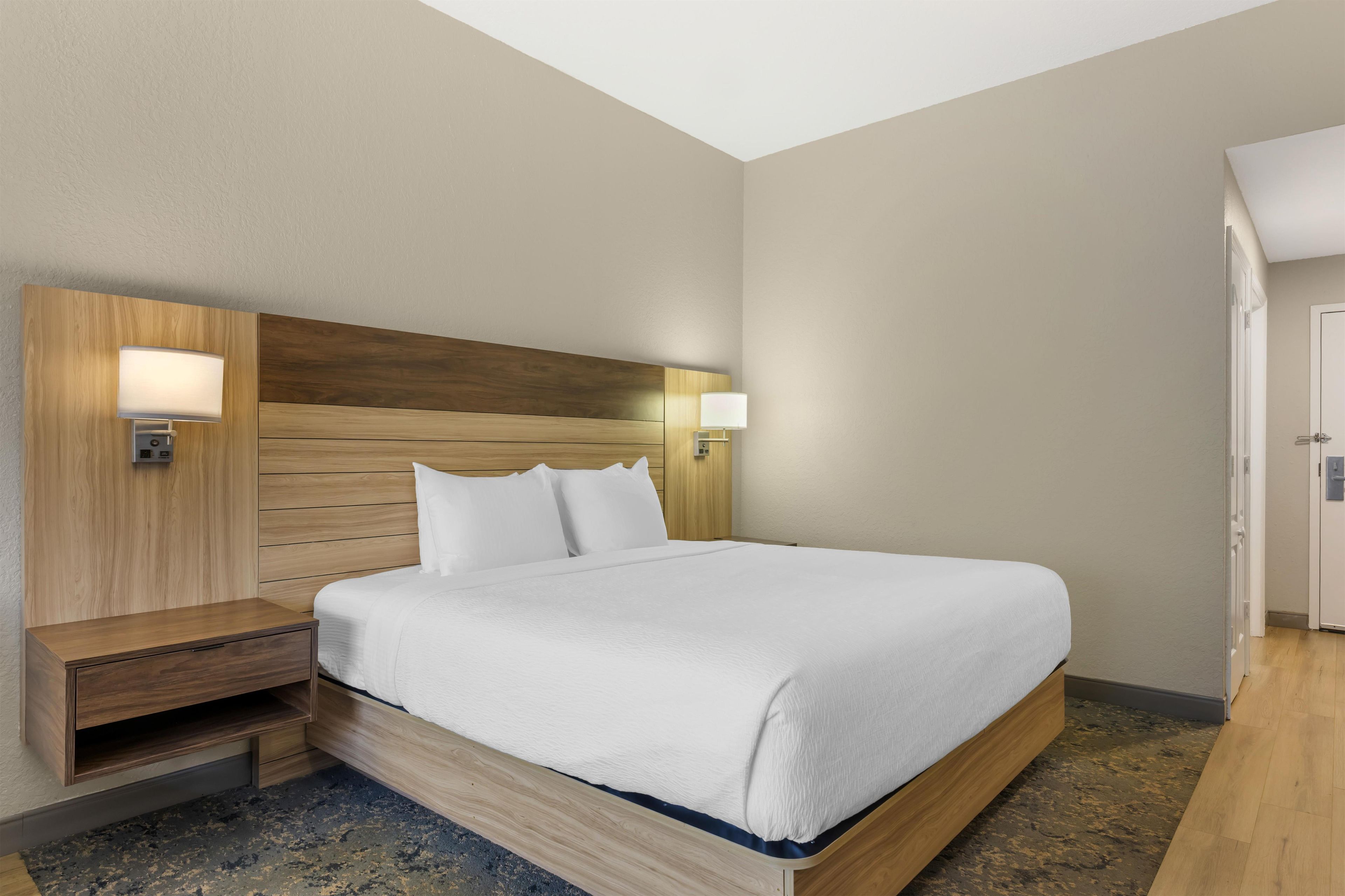 Una habitación de hotel con cabecera de madera, una cama con ropa de cama blanca, dos lámparas de noche y una mesita de noche de madera.