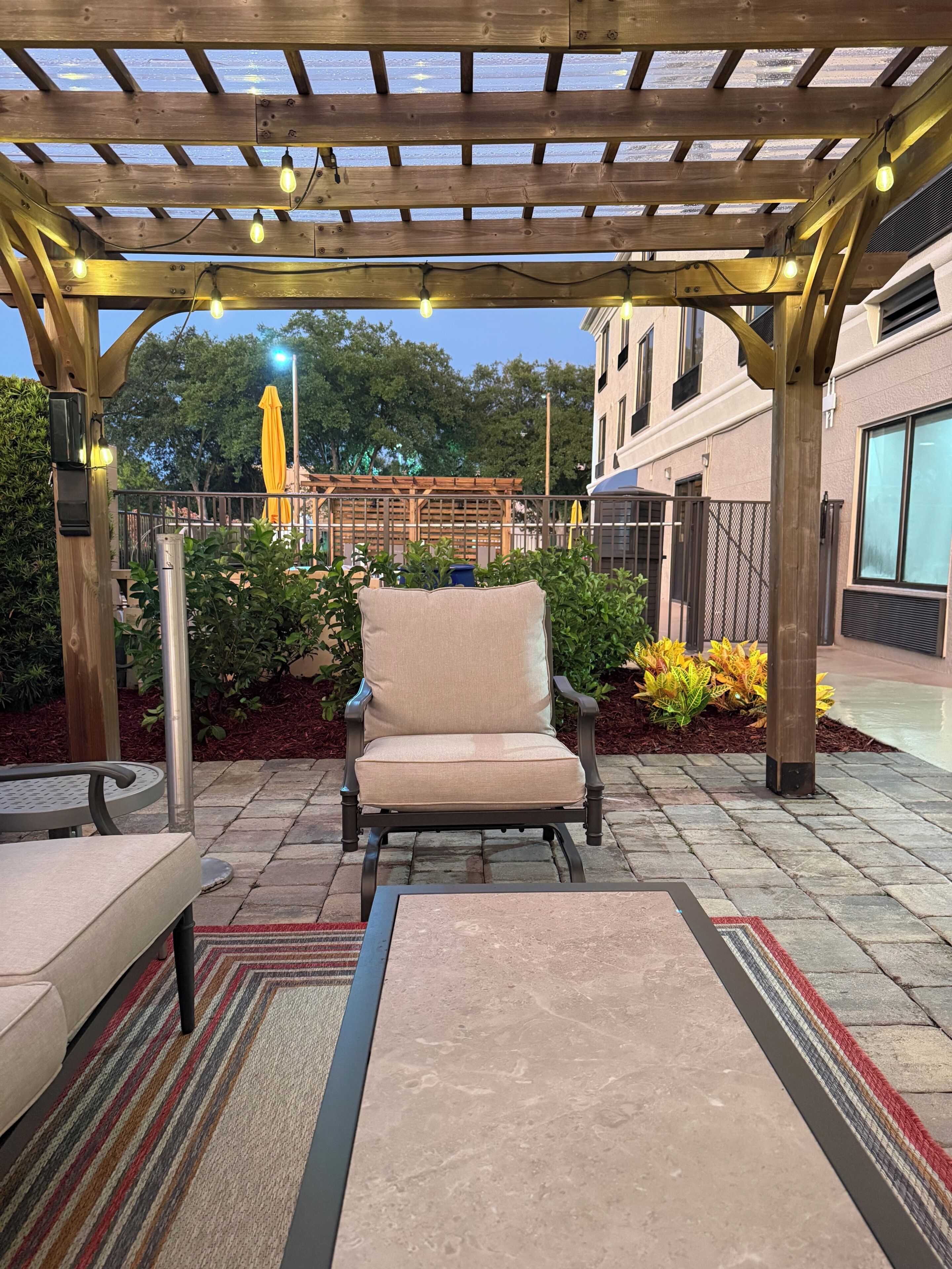 A covered patio area with a wooden pergola, a cushioned chair, and a table.
