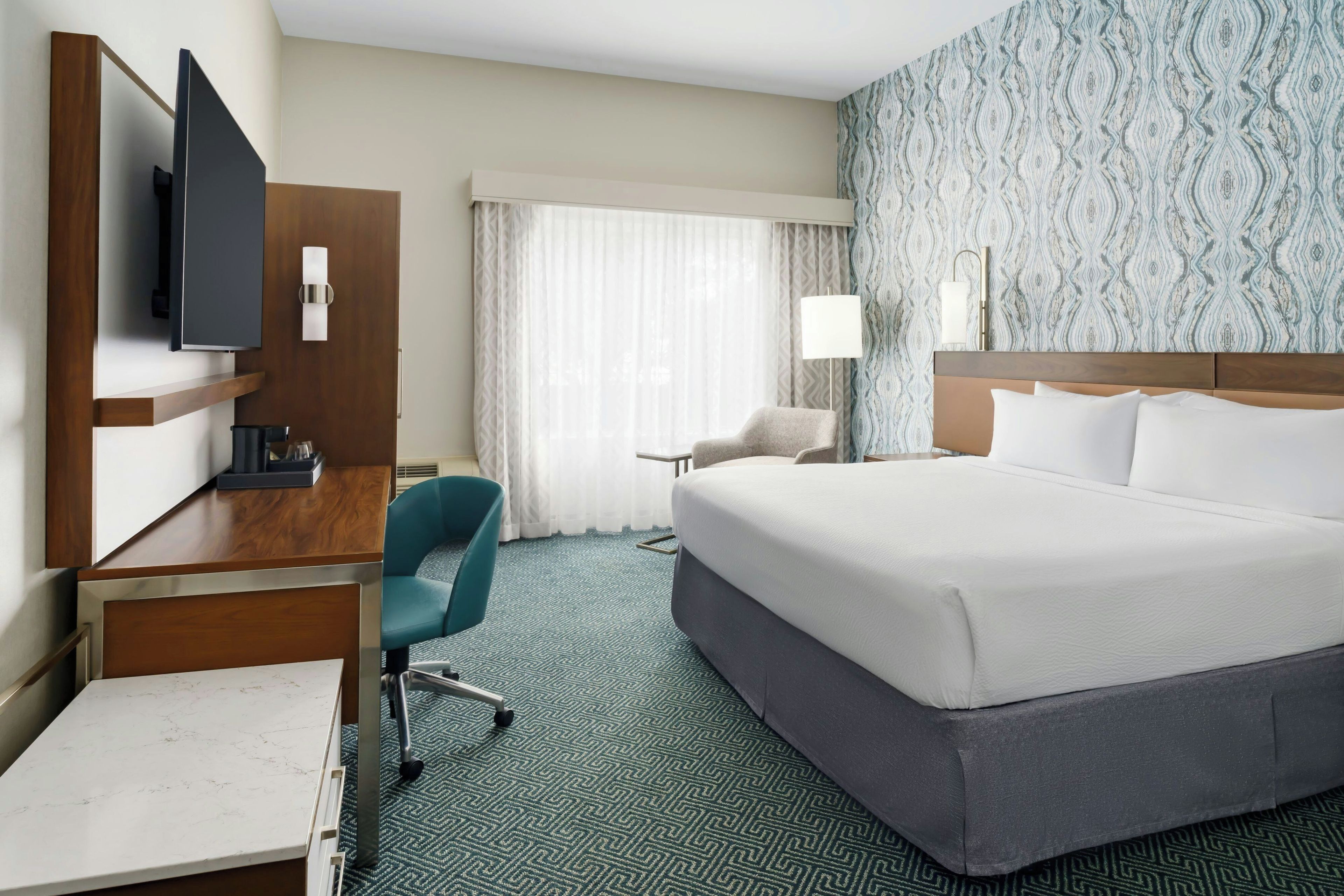Ein Hotelzimmer mit einem großen Bett, einem Schreibtisch mit Stuhl, einem Fernseher und einem Fenster mit Vorhängen.