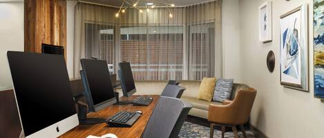 A conference room with two Apple iMacs, a keyboard, a mouse, a wooden desk, a grey office chair, a sofa with cushions, and a patterned rug.