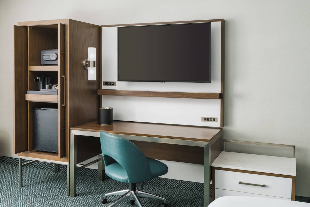 Ein Hotelzimmer mit Schreibtisch aus Holz, einem blaugrünen Bürostuhl, einem Flachbildfernseher, einer Minibar und einem kleinen weißen Nachttisch.