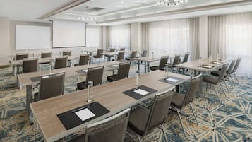 Un salón de conferencias con varias mesas y sillas, una pantalla de proyector y un amplio ventanal con cortinas.