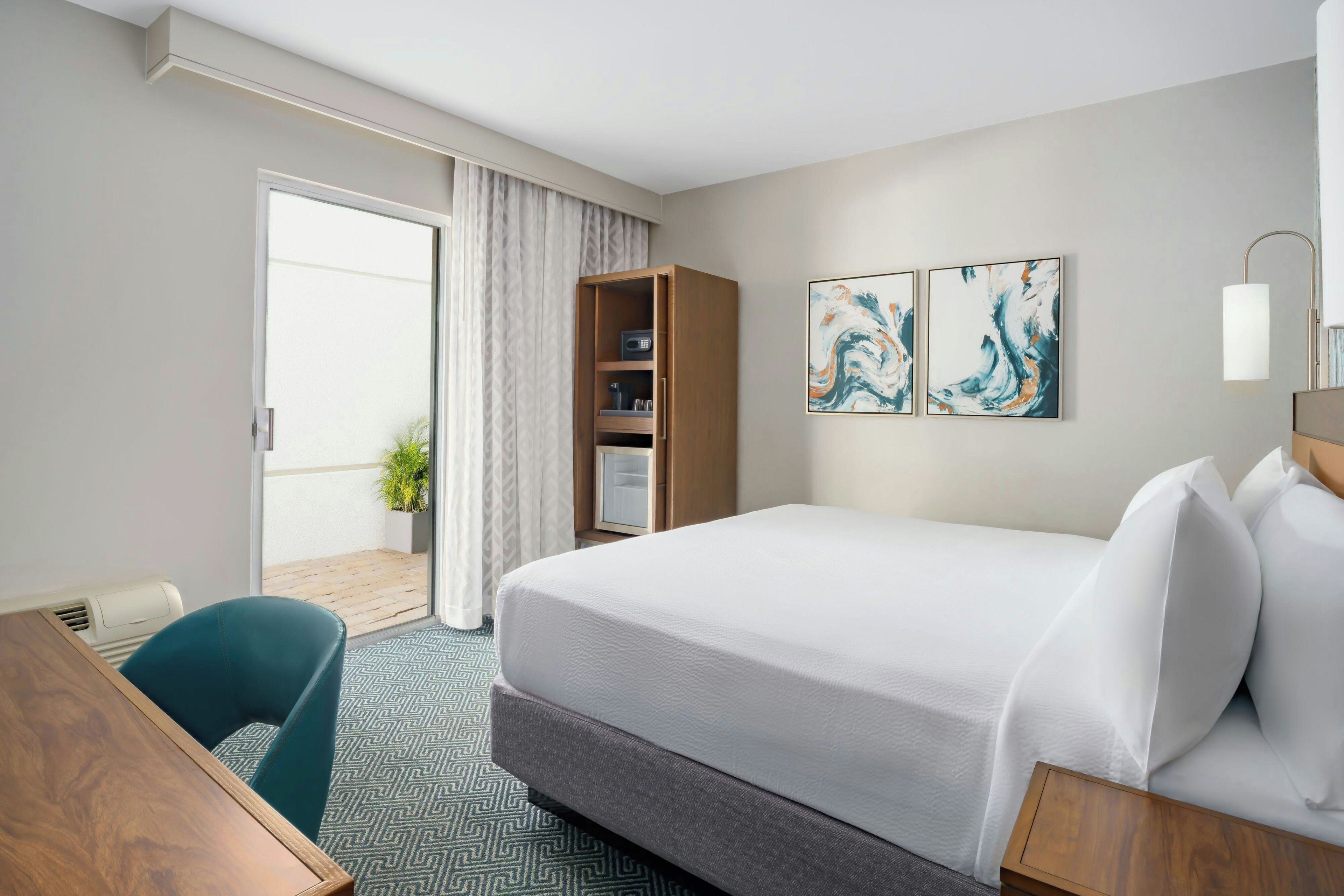 A hotel room with a bed, a desk, a chair, a wardrobe, and a small balcony with a plant.