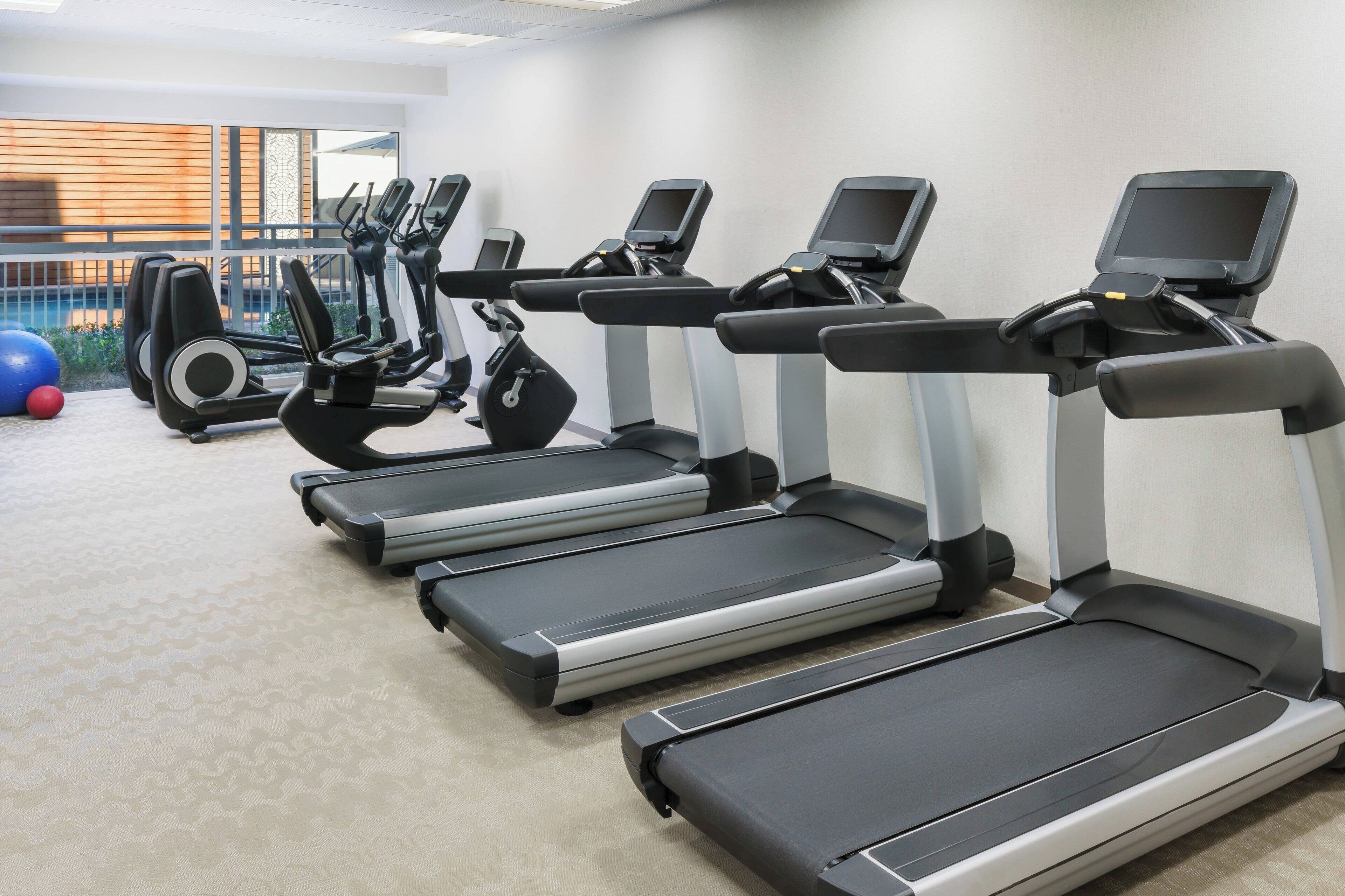 A row of treadmills in a gym with a patterned carpet and a view of a pool through large windows.