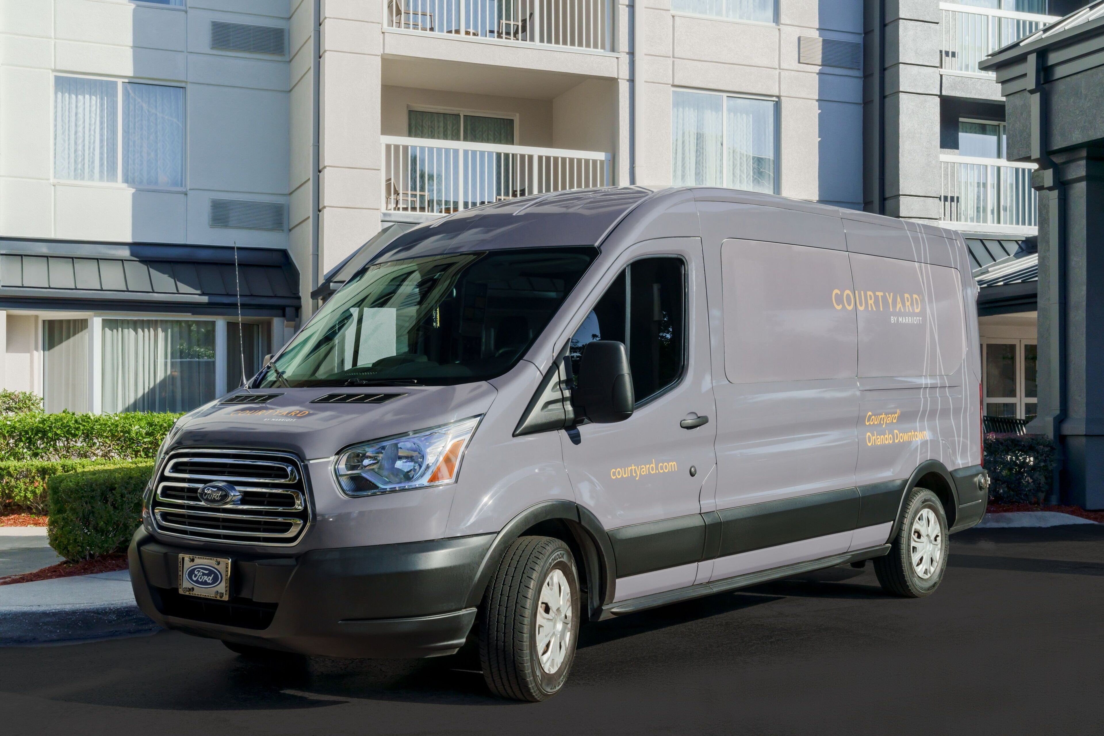 Een grijze Ford bestelwagen geparkeerd voor een hotel met balkons en grote ramen.