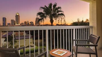 A balcony with wicker chairs and a table, overlooking a cityscape at sunset.