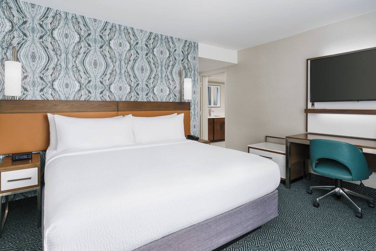 Ein Hotelzimmer mit einem großen Bett, einem Schreibtisch mit Stuhl, einem Fernseher und einer Wand mit Muster.