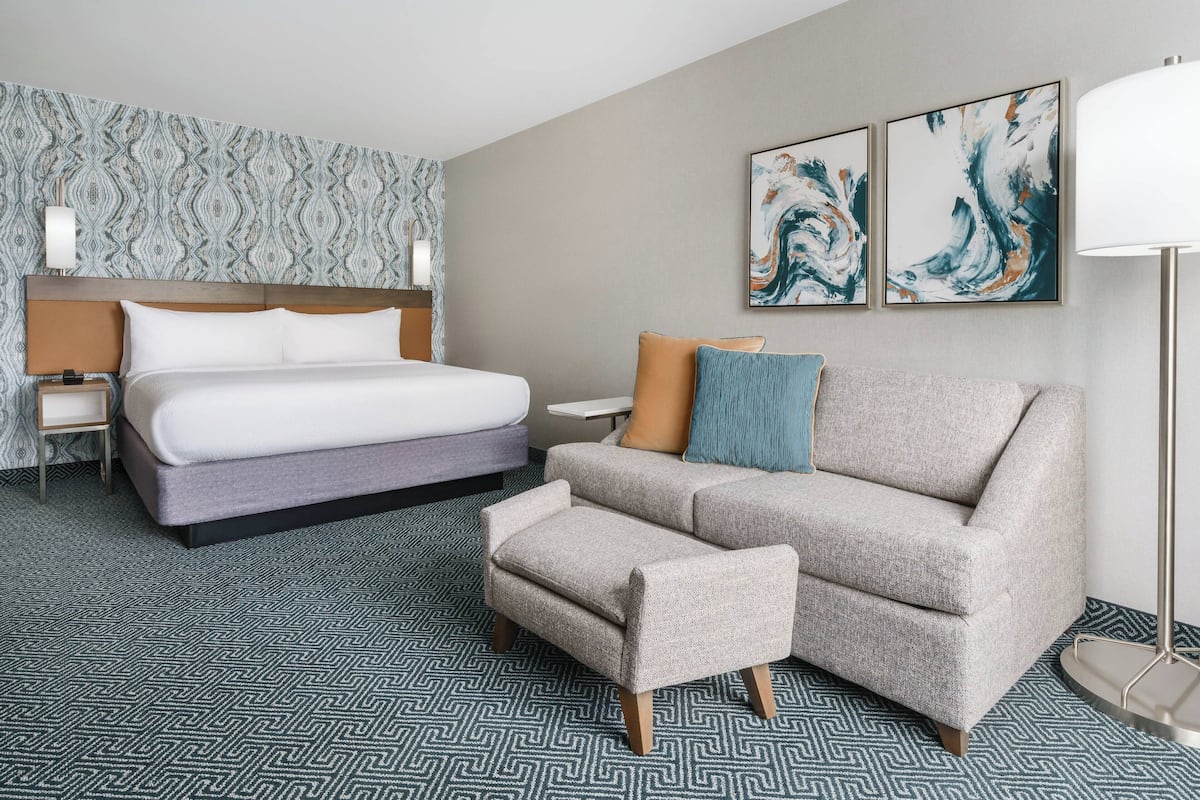 Ein Hotelzimmer mit Bett, Sofa, Hocker, Nachttisch, Lampe und abstrakten Wandbildern.
