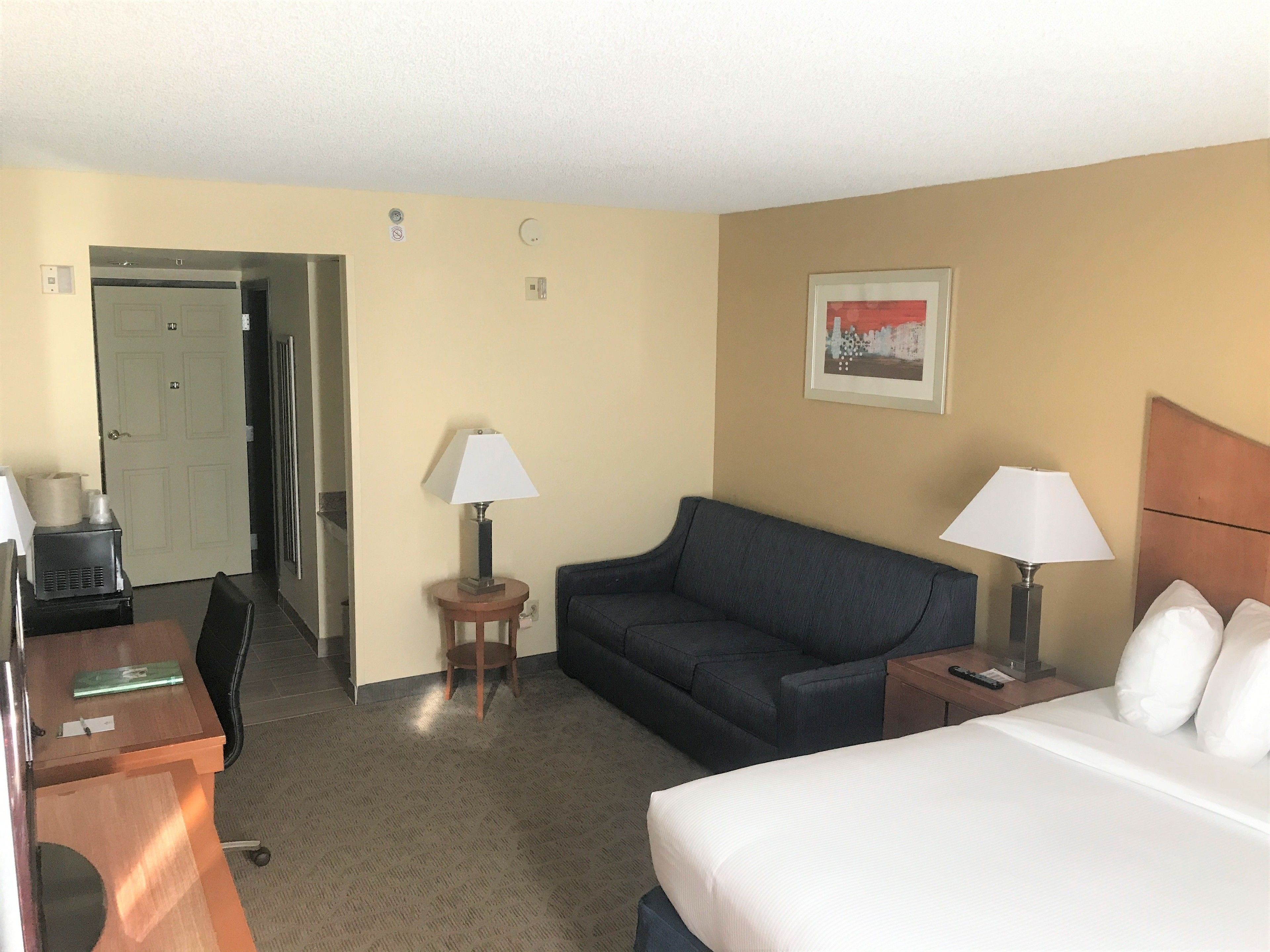 A hotel room with a bed, a sofa, a desk, and a chair. There is a door leading to another room and a small table with a lamp.