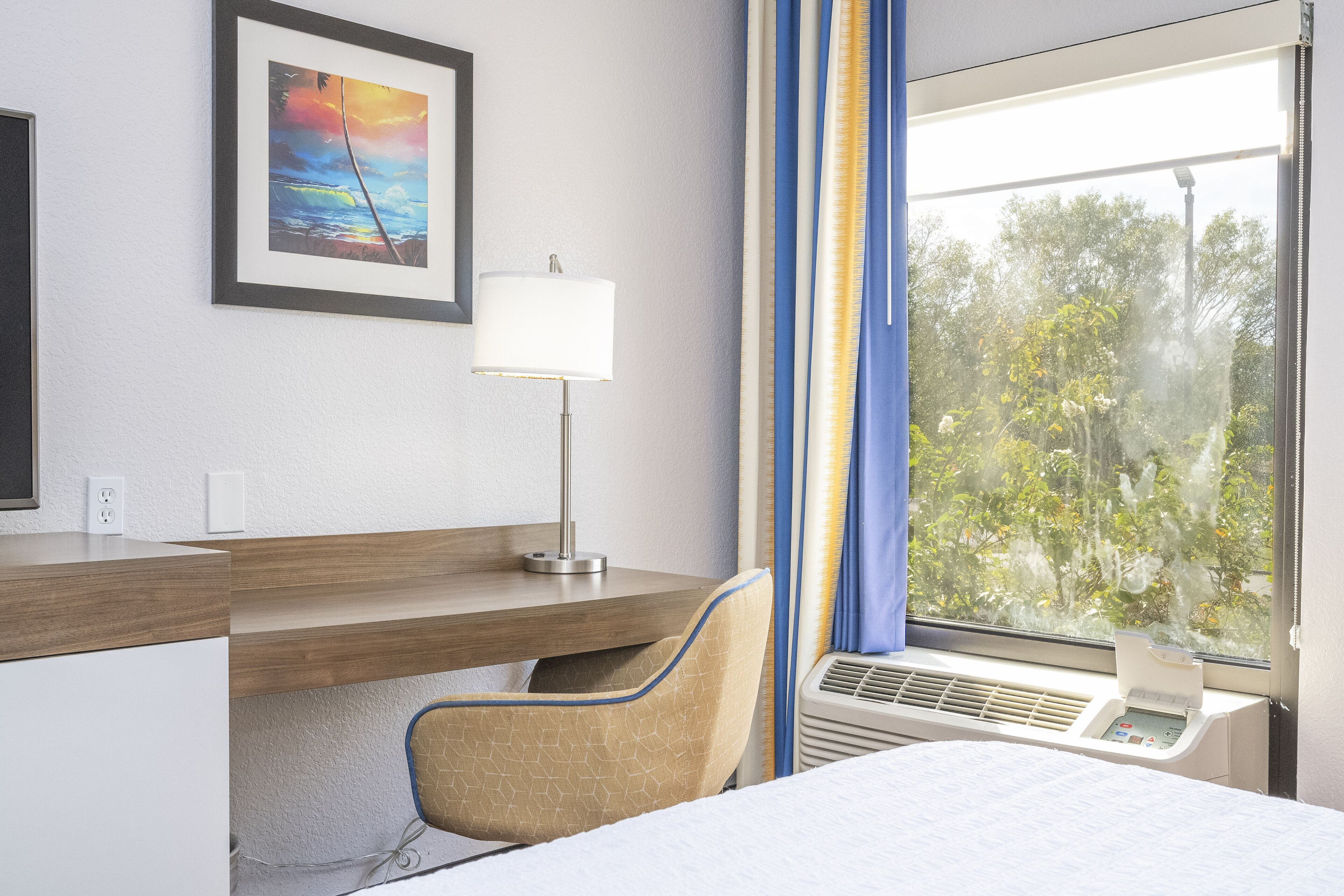 Une chambre d’hôtel comprenant un lit, un bureau avec une lampe, une chaise, une fenêtre avec des rideaux et un appareil de climatisation.