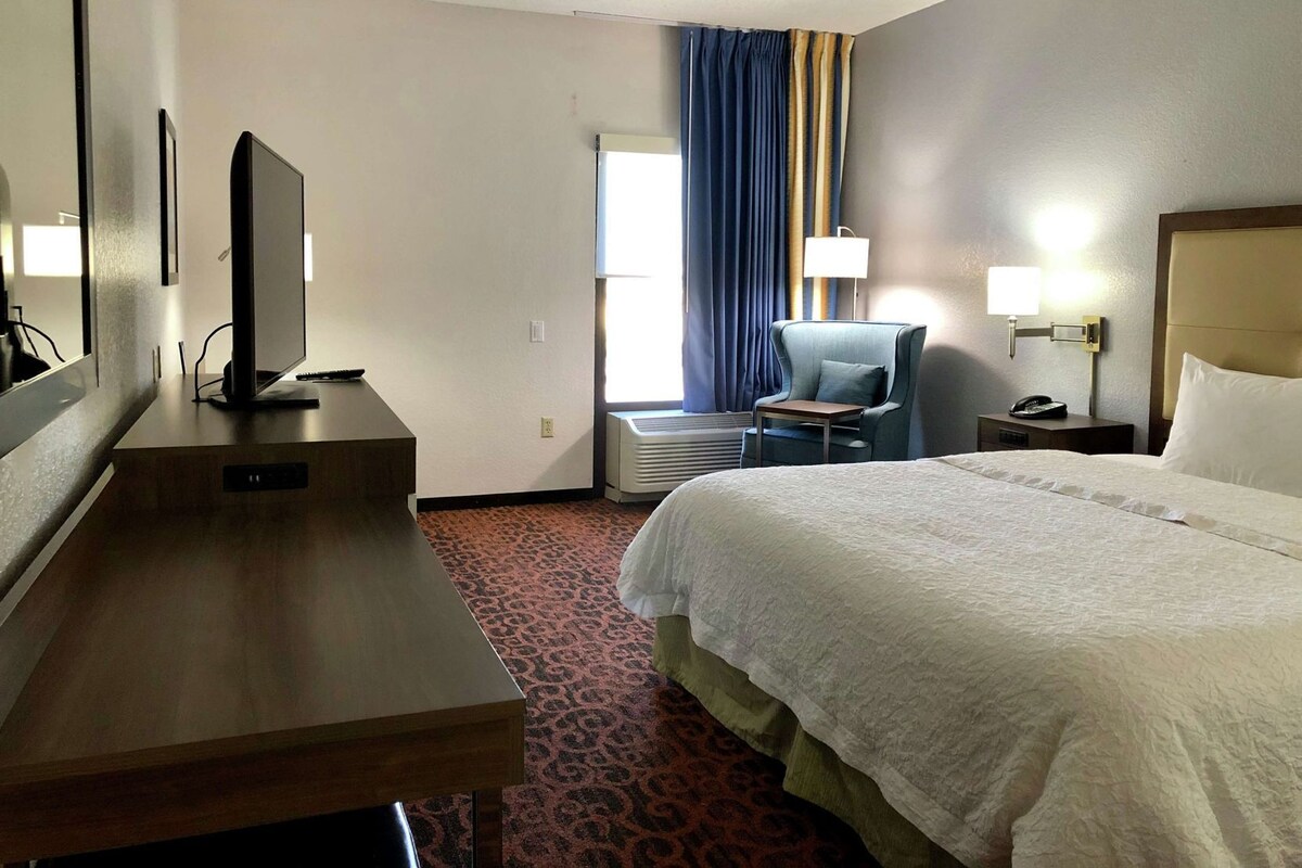 A hotel room with a bed, a desk, a chair, a TV, a lamp, and an air conditioning unit.