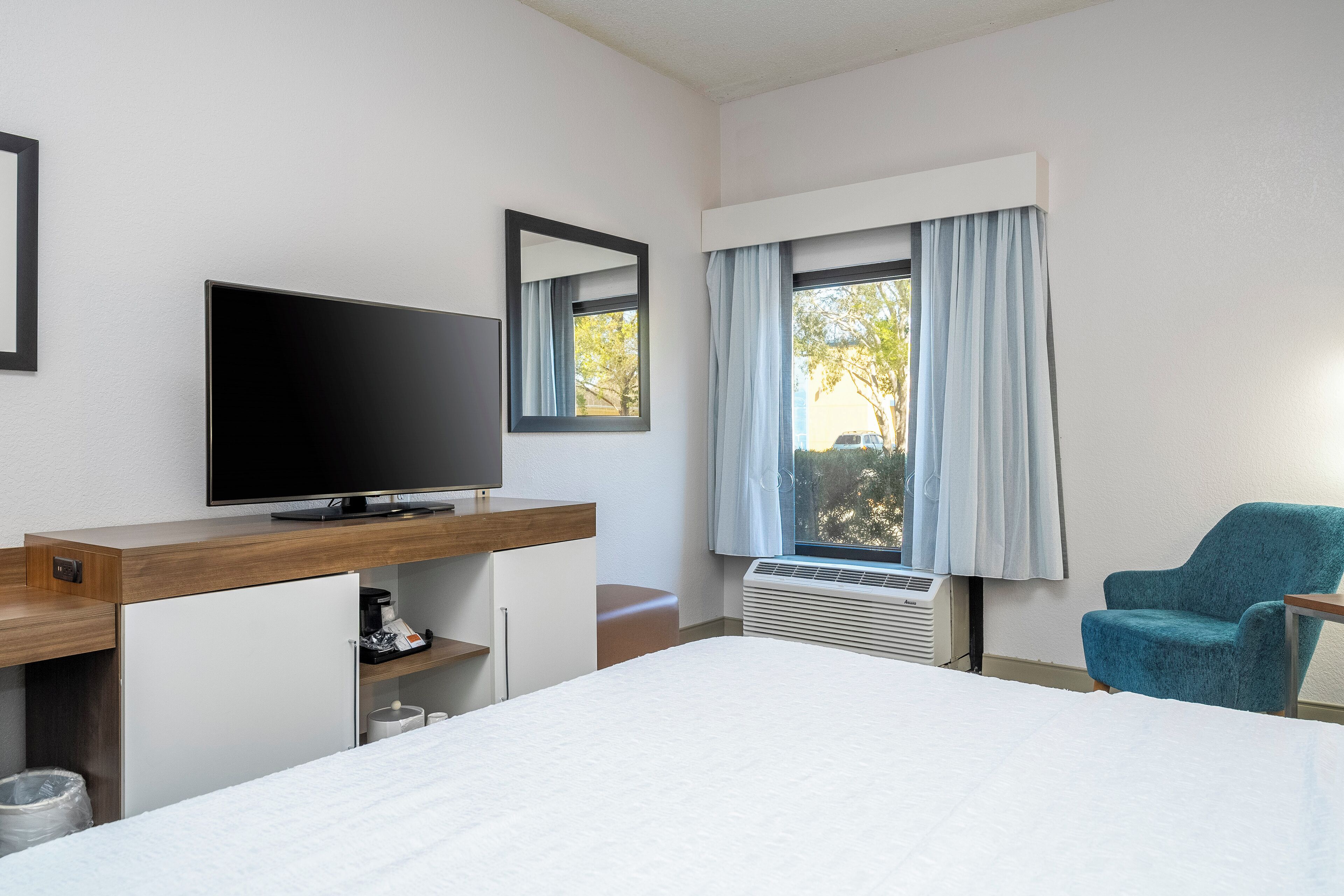 Habitación de hotel con cama, televisor, escritorio con computadora, silla y ventana con cortinas.