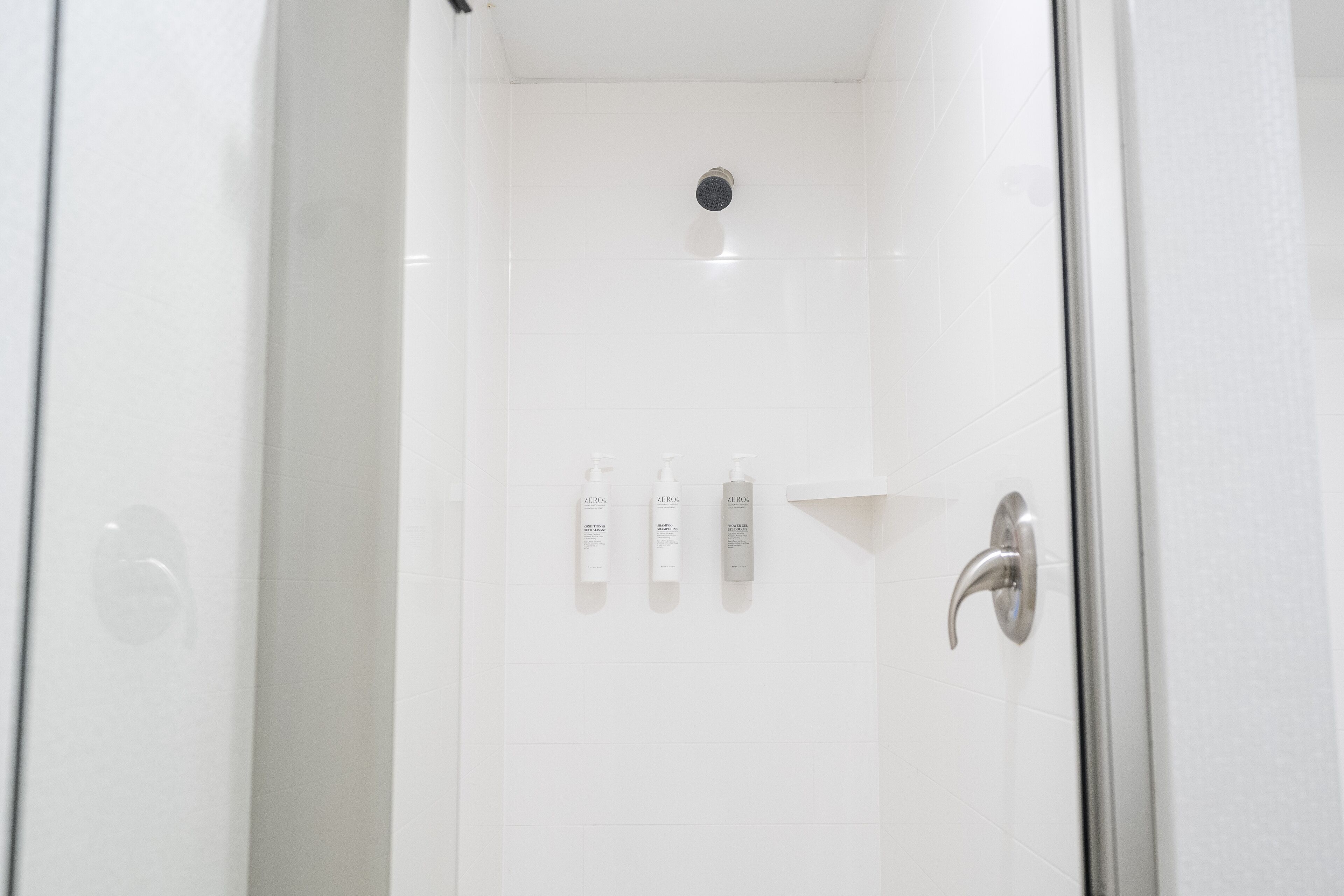 A white shower with a glass door, a showerhead, and three bottles of shampoo on the wall.