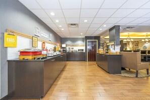 A hotel kitchen with a breakfast bar, a sign for eggs, and a seating area with a chair.