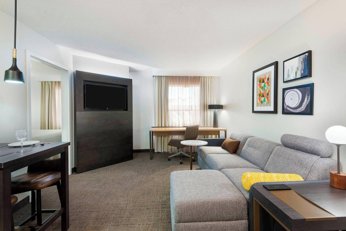 Una habitación de hotel moderna con un escritorio de madera oscura, un televisor de pantalla plana, un sofá gris y una mesa de comedor con sillas.