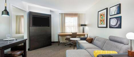 A modern hotel room with a dark wood desk, a flat-screen TV, a gray sofa, and a dining table with chairs.