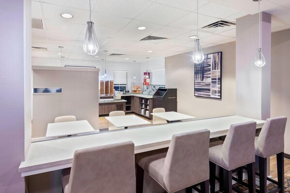 A modern conference room with a long table, several chairs, pendant lights, and a kitchen area in the background.