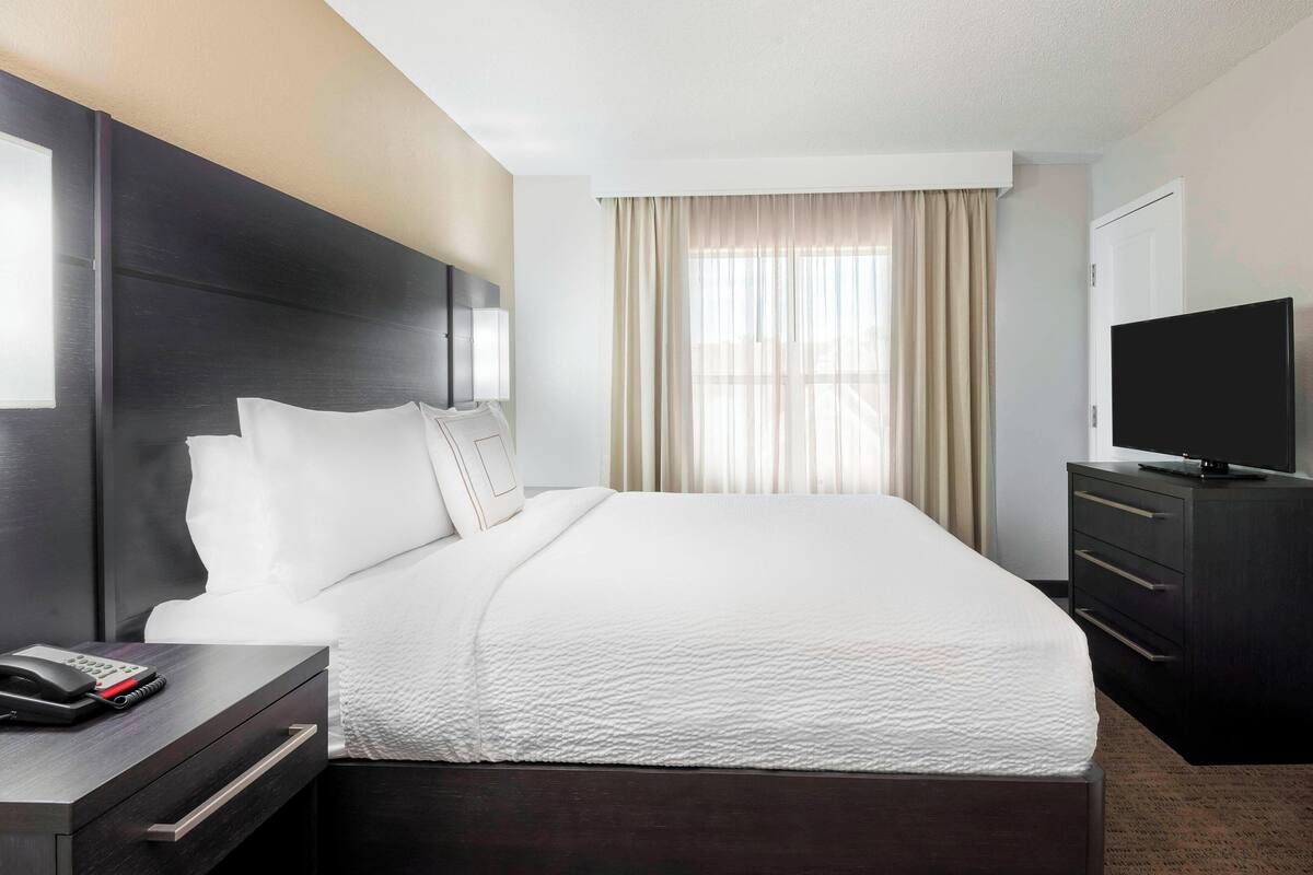 Habitación de hotel con una cama grande, una mesita de noche con teléfono, televisión y ventana con cortinas.