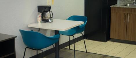 A small kitchenette with a coffee maker, two blue chairs, a white table, and a black refrigerator.