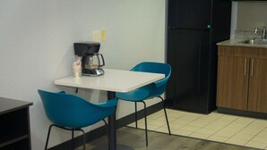 Eine kleine Küchenzeile mit einer Kaffeemaschine, zwei blauen Stühlen, einem weißen Tisch und einem schwarzen Kühlschrank.
