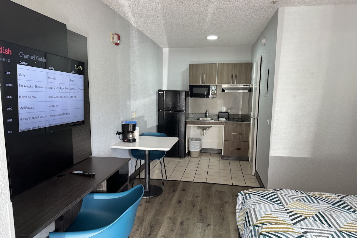A compact hotel room with a kitchenette, a dining area with a table and blue chairs, and a bed with a patterned bedspread.