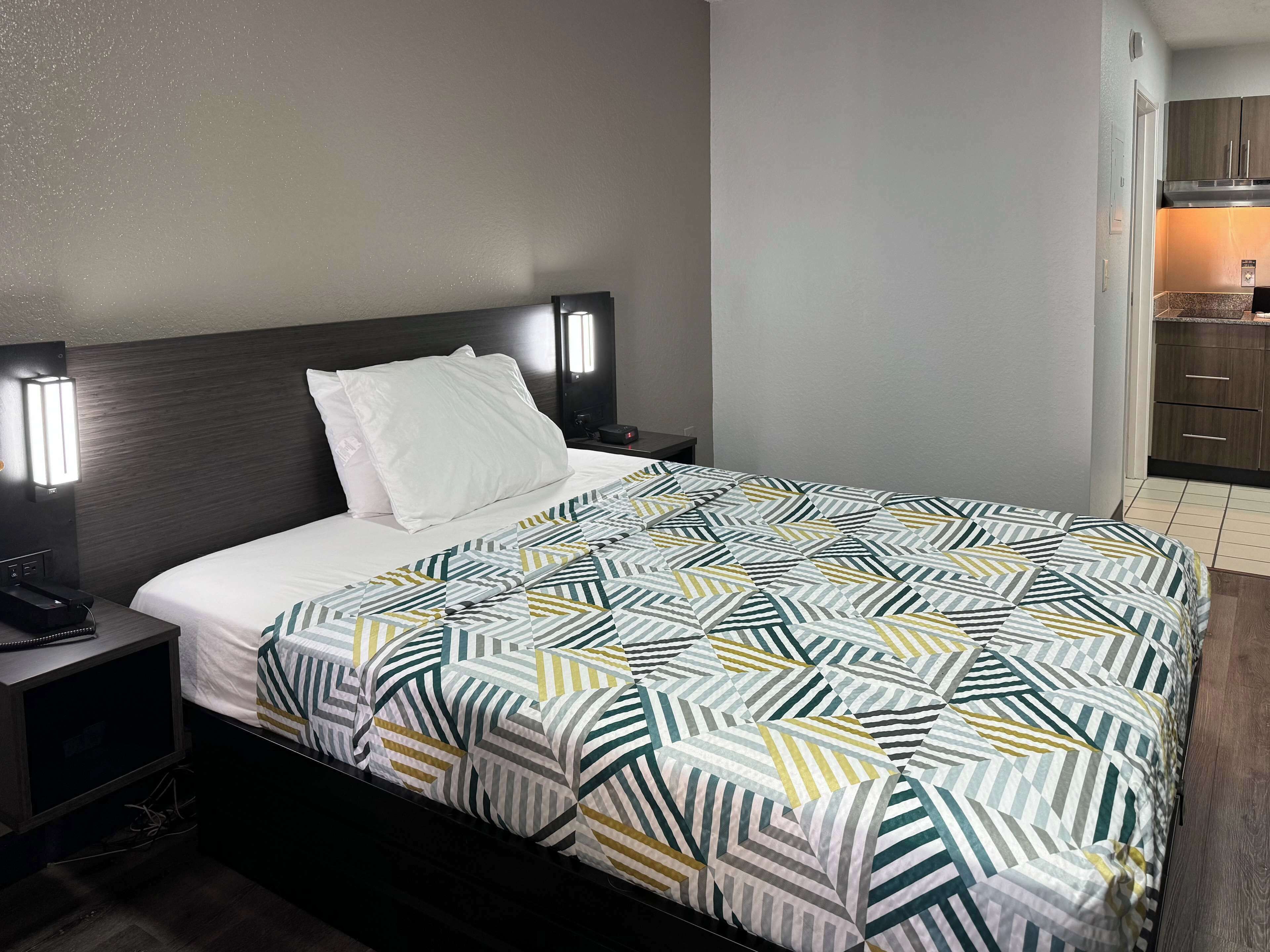 A hotel room with a neatly made bed, a bedside table with a phone, and a nightstand with a lamp.