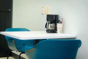 Een kleine tafel met een koffiezetapparaat en een kop koffie. Er staan twee blauwe stoelen bij de tafel.