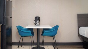Ein kleines Zimmer mit einem weißen Tisch, zwei blauen Stühlen, einer Kaffeemaschine, einem Bett und einem Kühlschrank.