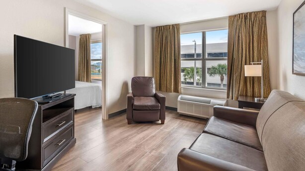 A hotel room with a TV, a leather chair, a sofa, a desk with a lamp, and a large window with curtains.